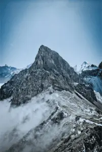 A rocky mountain peak with white snow and clouds nearby.